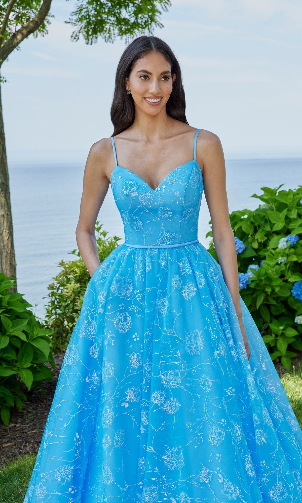 A woman stands outdoors by greenery and water, wearing the Morilee 49153 long prom dress—a light blue, sleeveless, floral-patterned A-line gown with thin straps.
